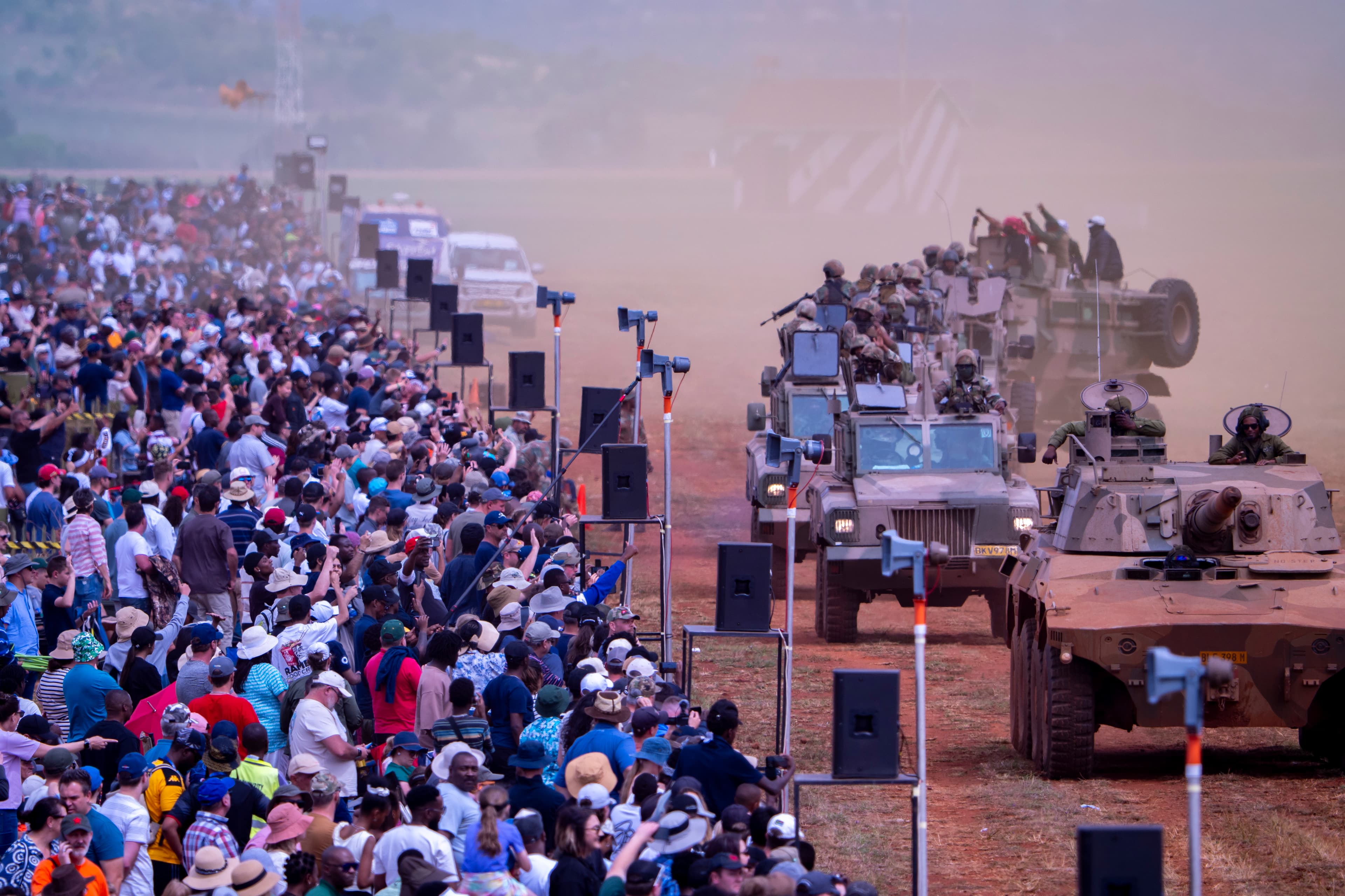 A large crowd watches military armored vehicles and tanks driving through a dusty field.